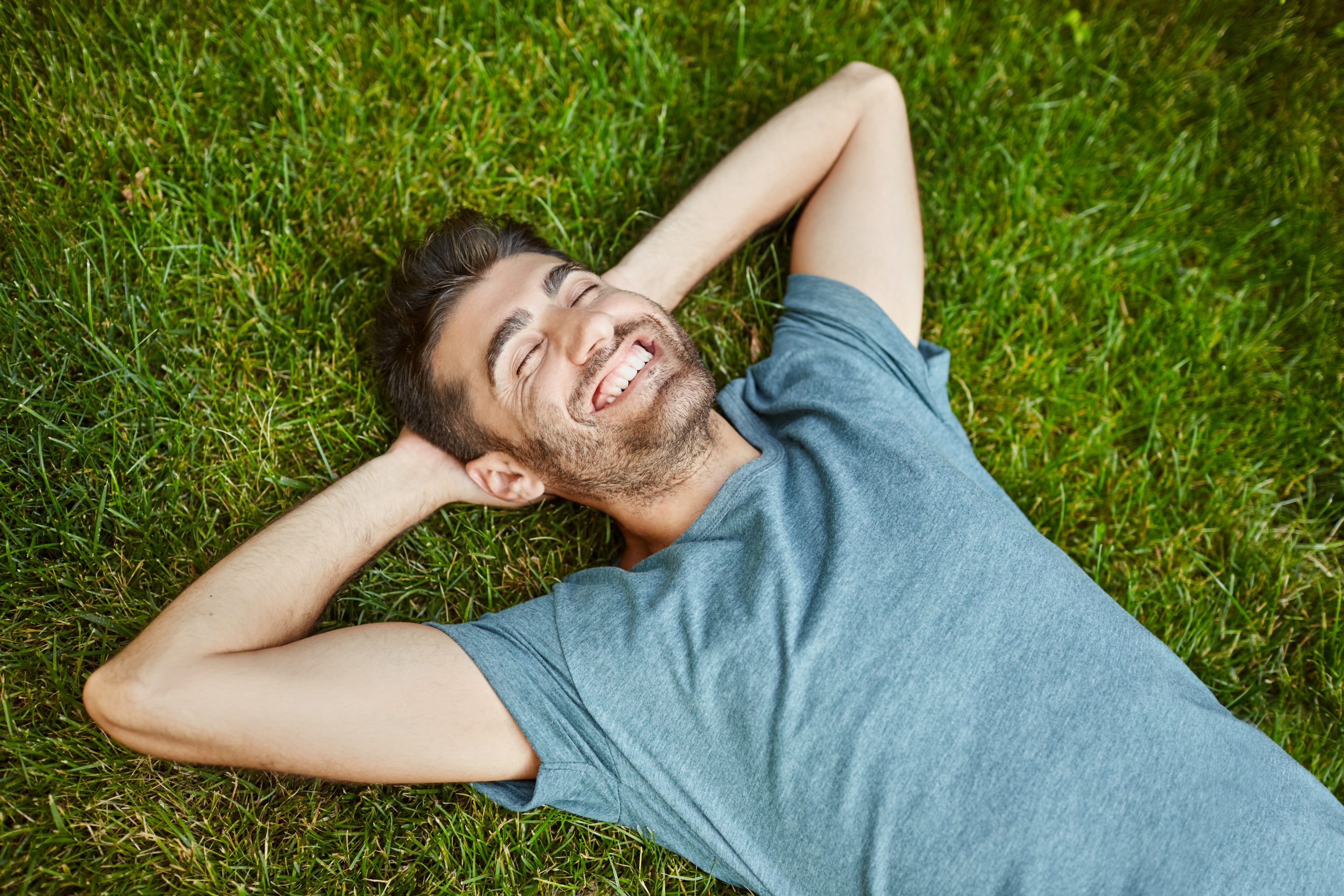 positive emotions young beautiful bearded caucasian male in blue t shirt lying on grass smiling with teeth laughing relaxing outside in summer morning with happy face expression scaled